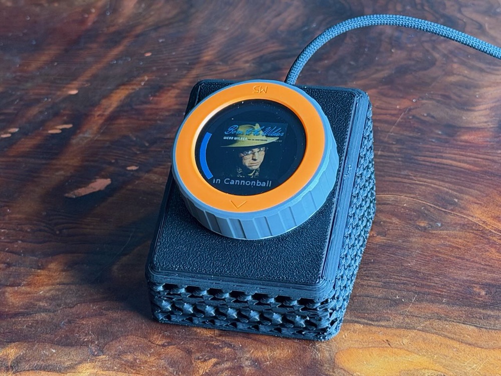 The M5Stack Dial in a black 3D-printed sloped case on a wooden desk. The round screen shows album artwork for Webb Wilder's Born To Be Wilder, a blue volume arc up the left side, and a scrolling track title along the bottom. A blue braided USB-C cable runs out of the back.